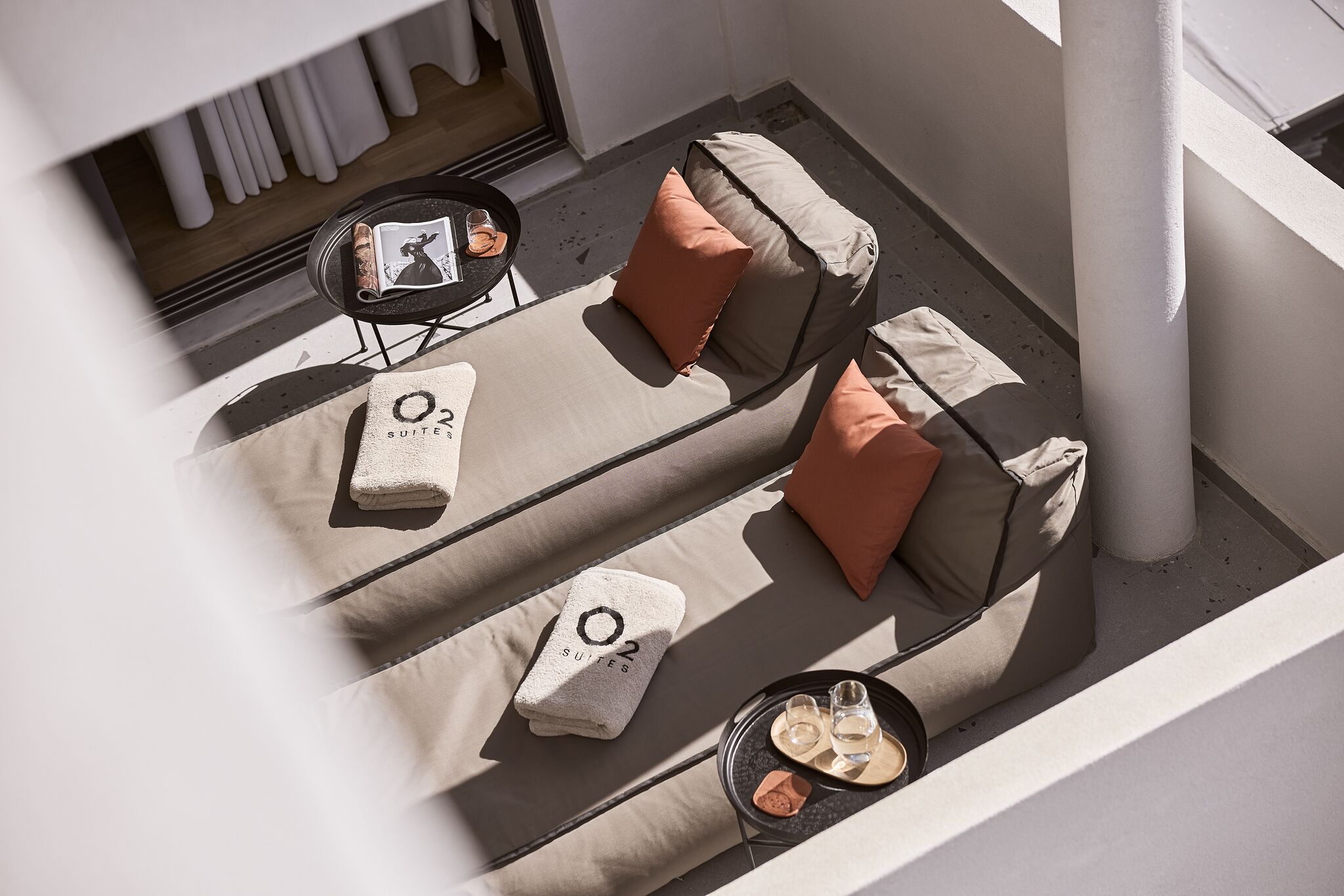 Un área de estar al aire libre iluminada por el sol, con dos sofás acolchados, una pequeña mesa con bebidas y una toalla con la marca 'O2 SUITES'.