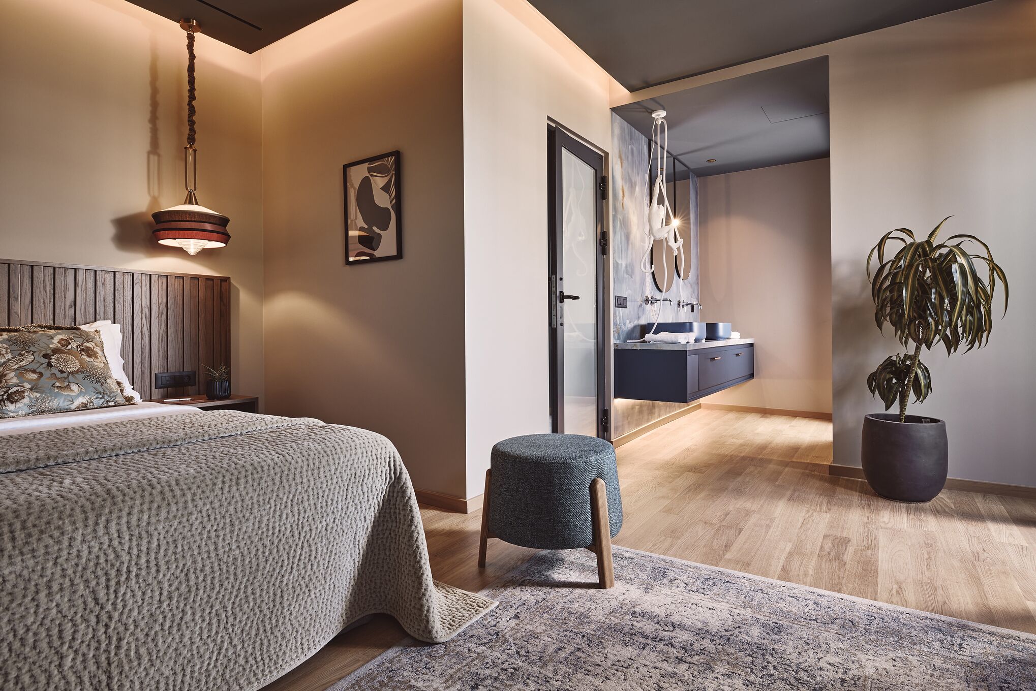 Un dormitorio moderno con cama, un taburete pequeño, una planta en maceta y un baño visible a través de una puerta abierta.