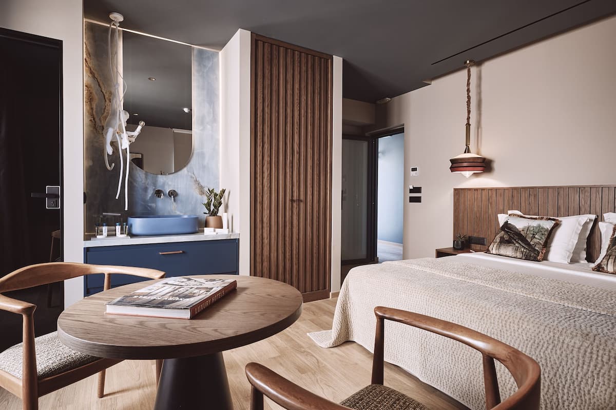 A modern hotel room with a large bed, a round wooden table with a book, a blue sink, and a wooden panel wall.