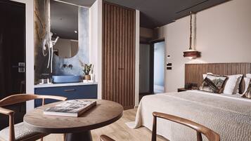 A modern hotel room with a large bed, a round wooden table with a book, a blue sink, and a wooden panel wall.