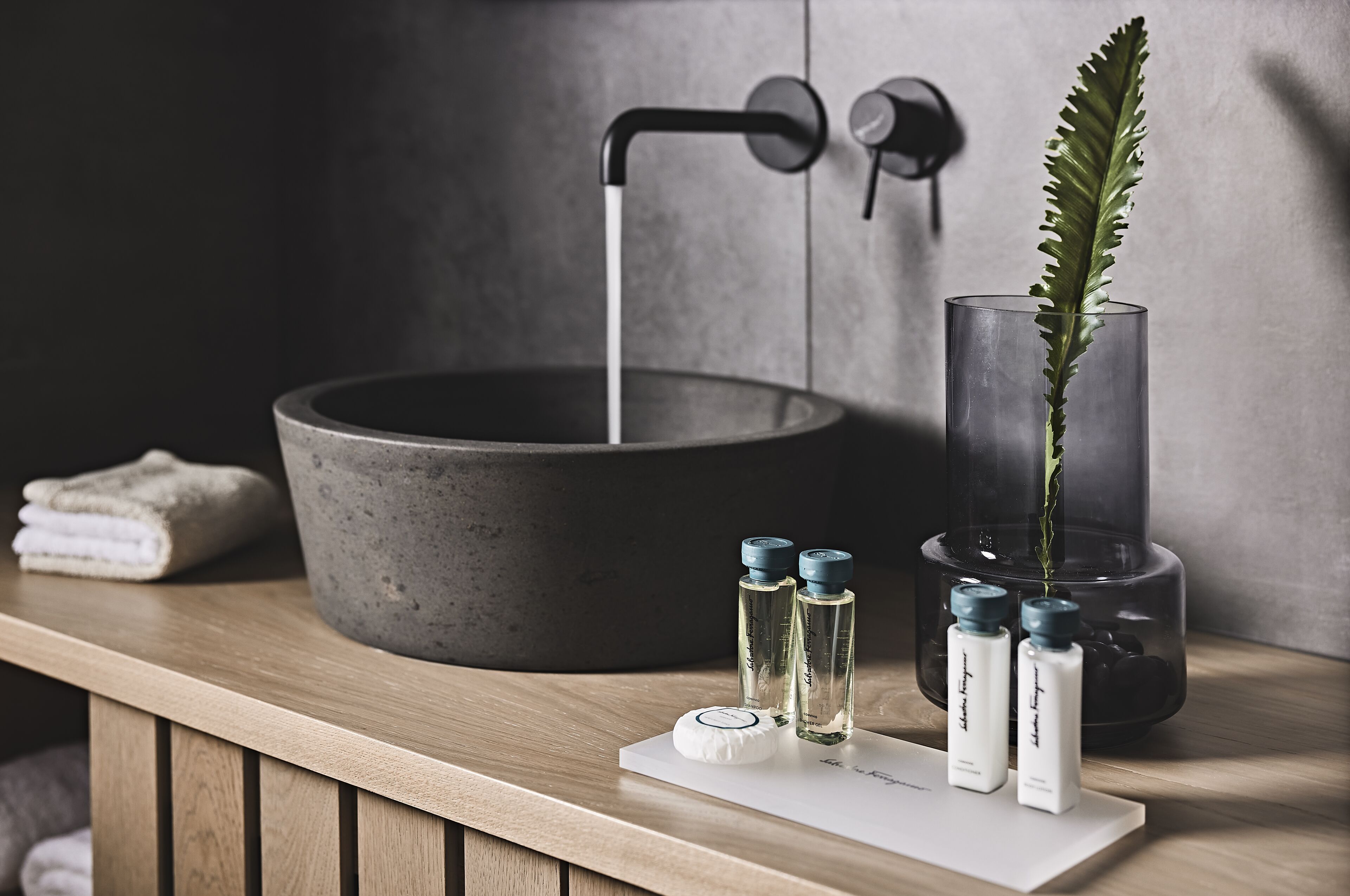 A bathroom with a large basin, a faucet, and a glass vase with a plant. There are also two bottles and a rolled towel on the counter.