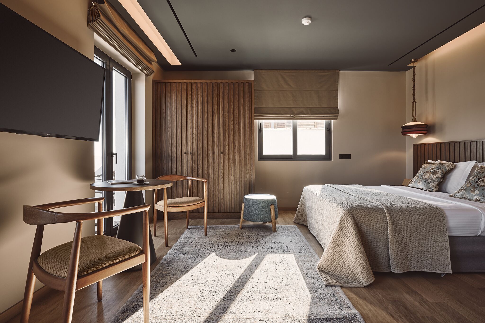 A modern hotel room with a large bed, a desk with a chair, a small stool, and a TV mounted on the wall.