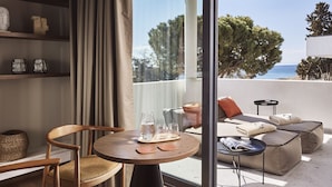 Una sala de estar moderna con una silla de madera, una mesa redonda con vasos y un sofá con cojines. Un ventanal grande ofrece vistas a los árboles y al mar.
