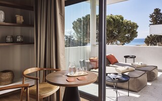 A modern living room with a wooden chair, a round table with glasses, and a sofa with cushions. A large window offers a view of trees and the sea.