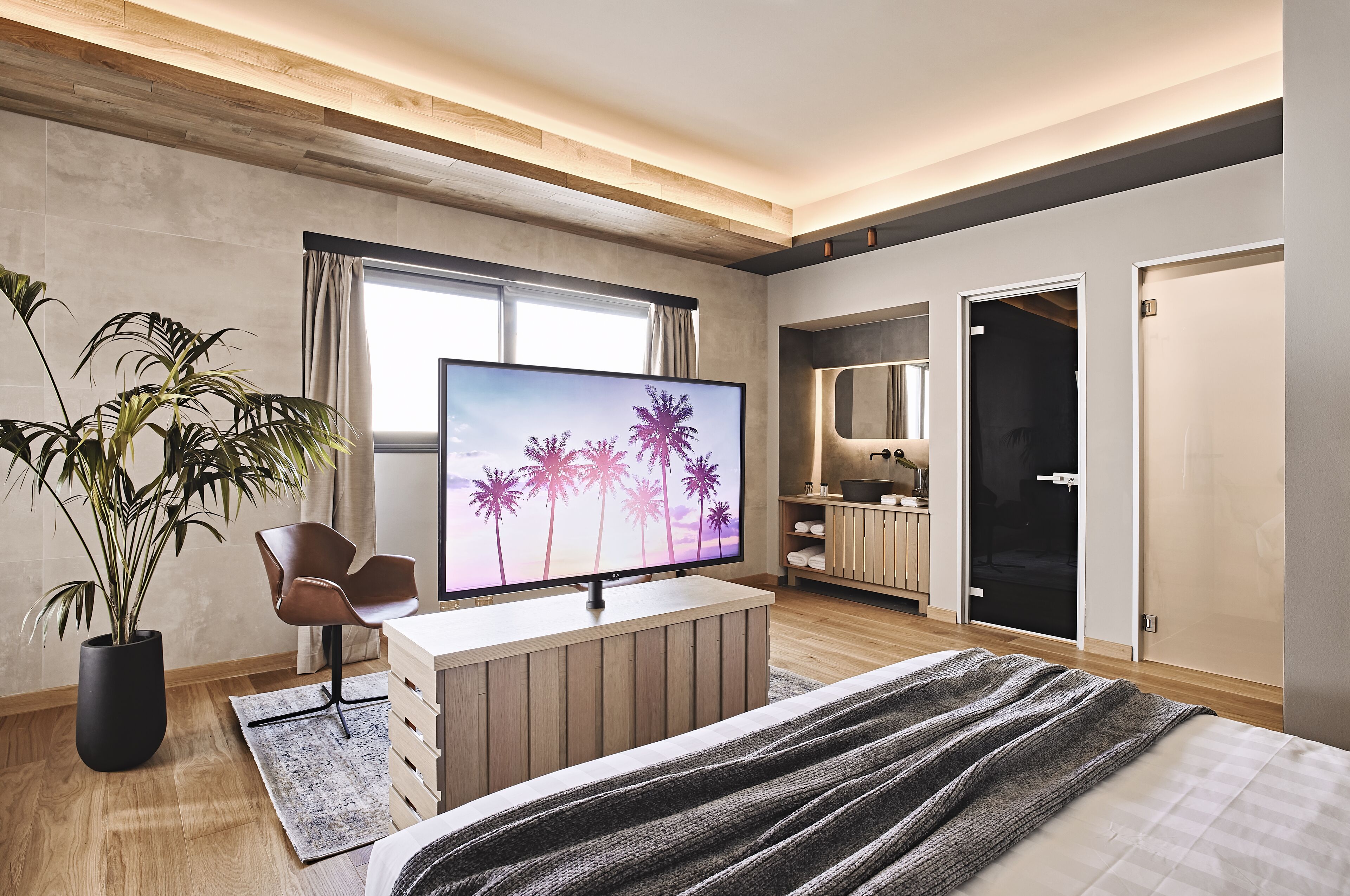 A modern hotel room with a large flat-screen TV, a wooden desk, a comfortable chair, a bed with a striped blanket, a potted plant, and a large mirror.