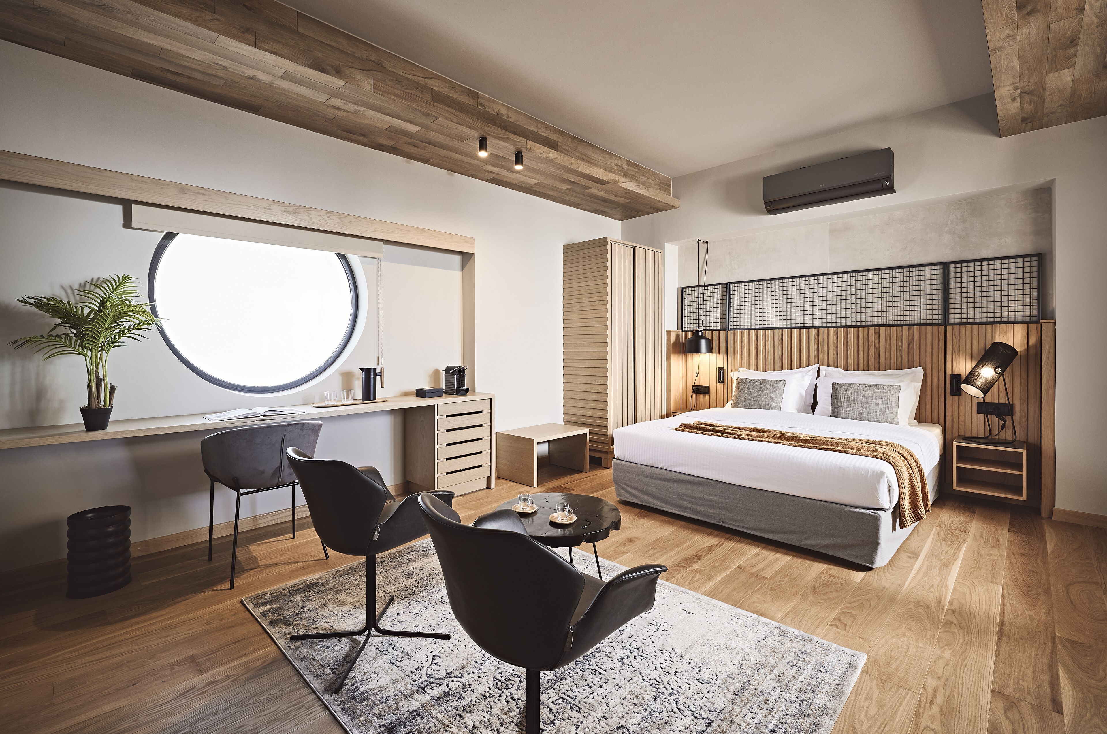 A modern bedroom with a large bed, a desk, and a chair, featuring wooden flooring and a large window.