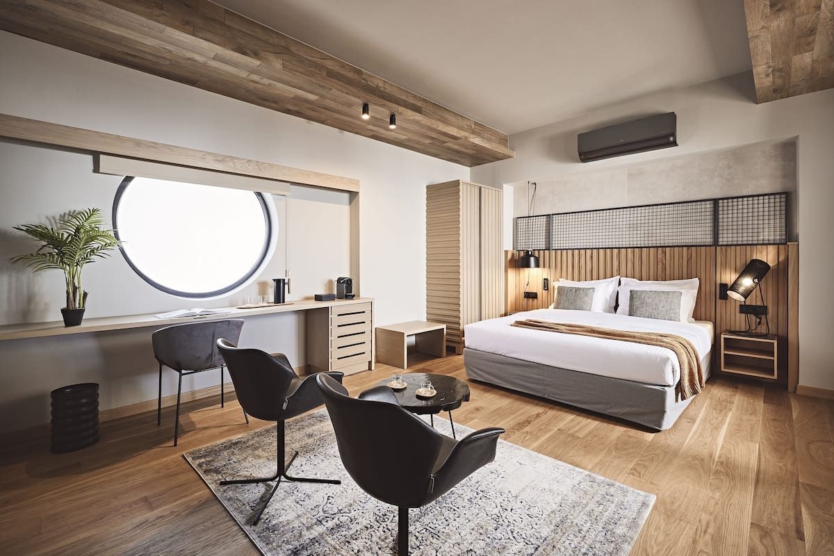 A modern bedroom with a large bed, a desk, a chair, a round window, and a potted plant.