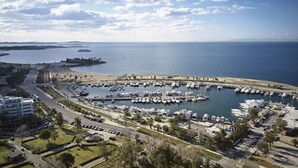 Un puerto deportivo con barcos amarrados, una carretera que conduce al puerto y una playa al fondo.
