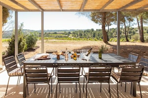 Un área de comedor al aire libre con una mesa preparada para seis personas, rodeada de sillas, con vistas a un viñedo y un paisaje montañoso.