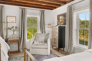 Ein Schlafzimmer mit Bett, Stuhl, Kommode, Lampe und einem Fenster mit Blick auf Grünflächen.