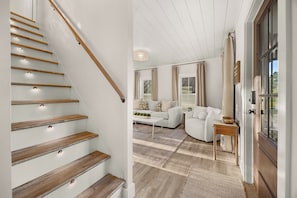 A staircase with wooden steps and a light fixture, leading to a living room with a sofa, a coffee table, and a view through a window.