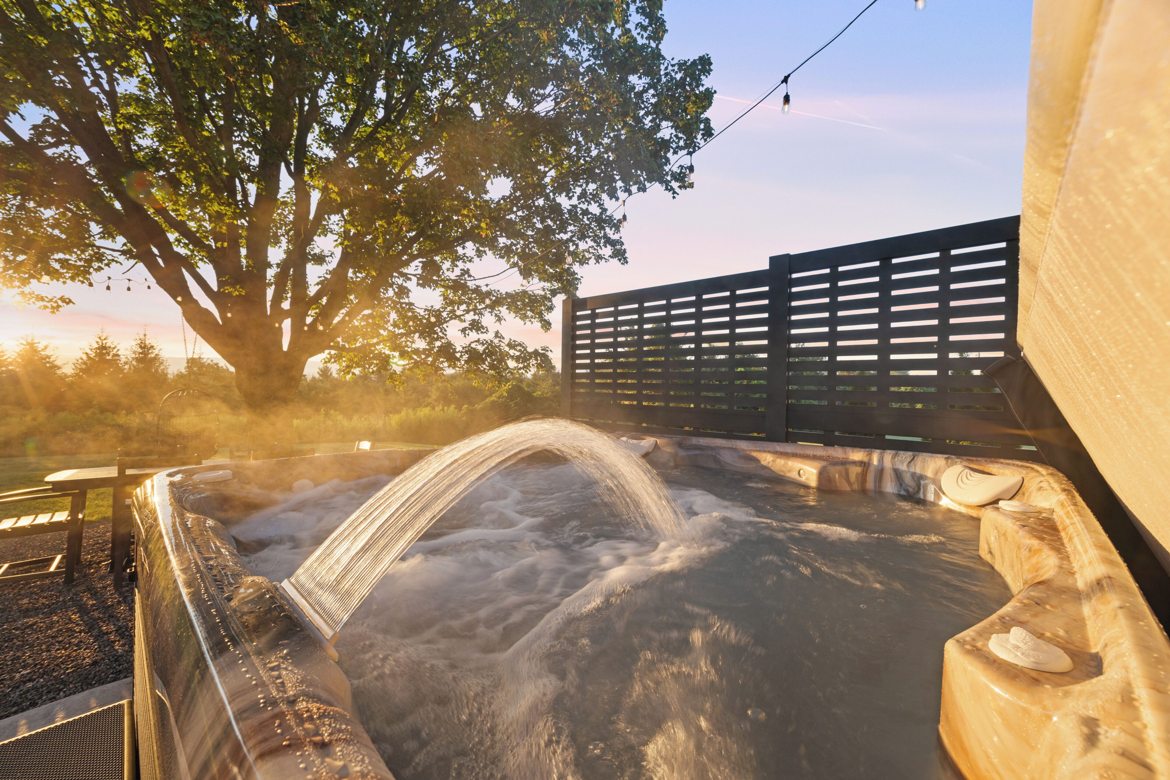 A hot tub with a waterfall feature, surrounded by trees and a wooden fence.