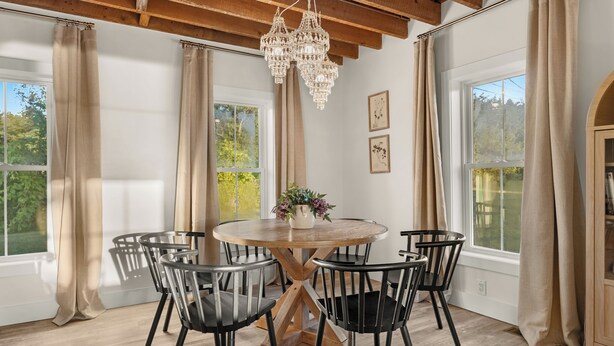 A dining area with a round table, six chairs, a chandelier, and large windows with curtains.