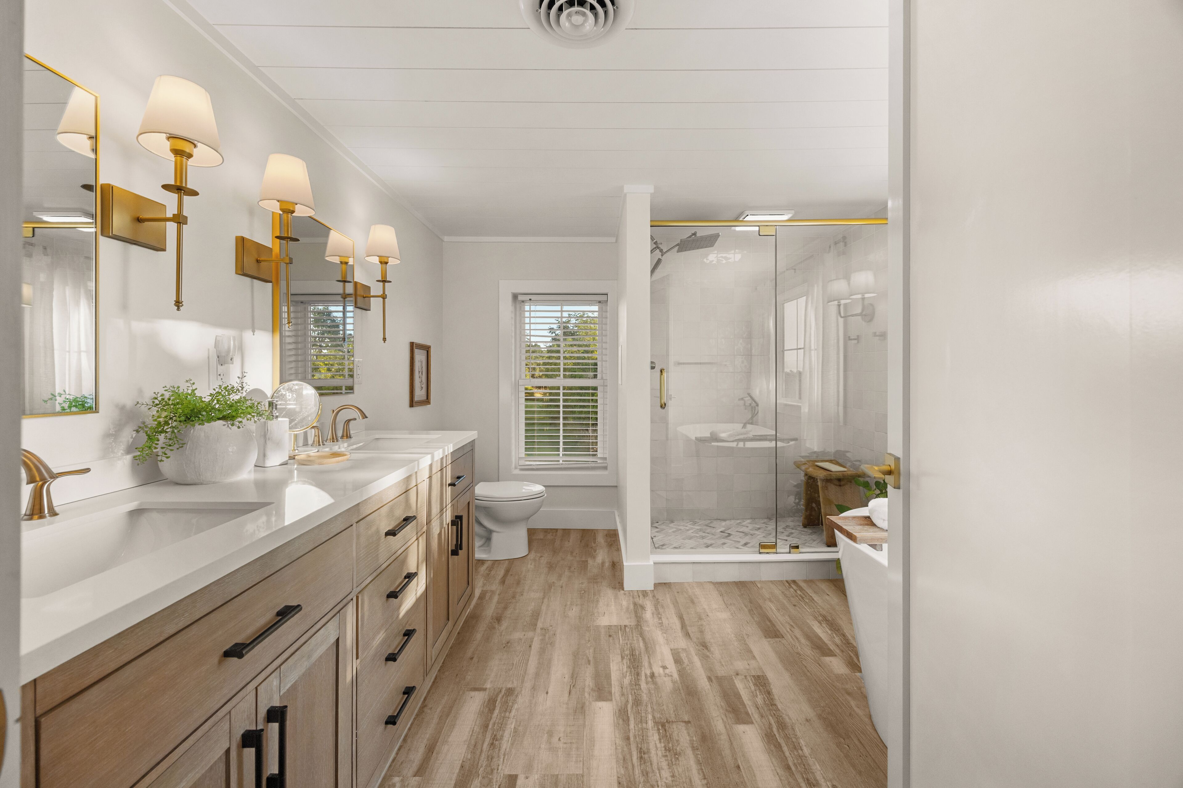 A modern bathroom with a double vanity, a toilet, a shower area, and a window with blinds.