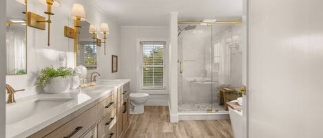 A modern bathroom with a double vanity, a toilet, a shower area, and a window with blinds.