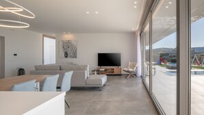 Una sala de estar moderna con sofá, televisor y comedor, ventanas amplias con vista a una piscina y montañas.