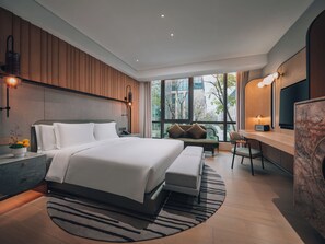 A modern hotel room with a large bed, a desk, and a view of the city through large windows.