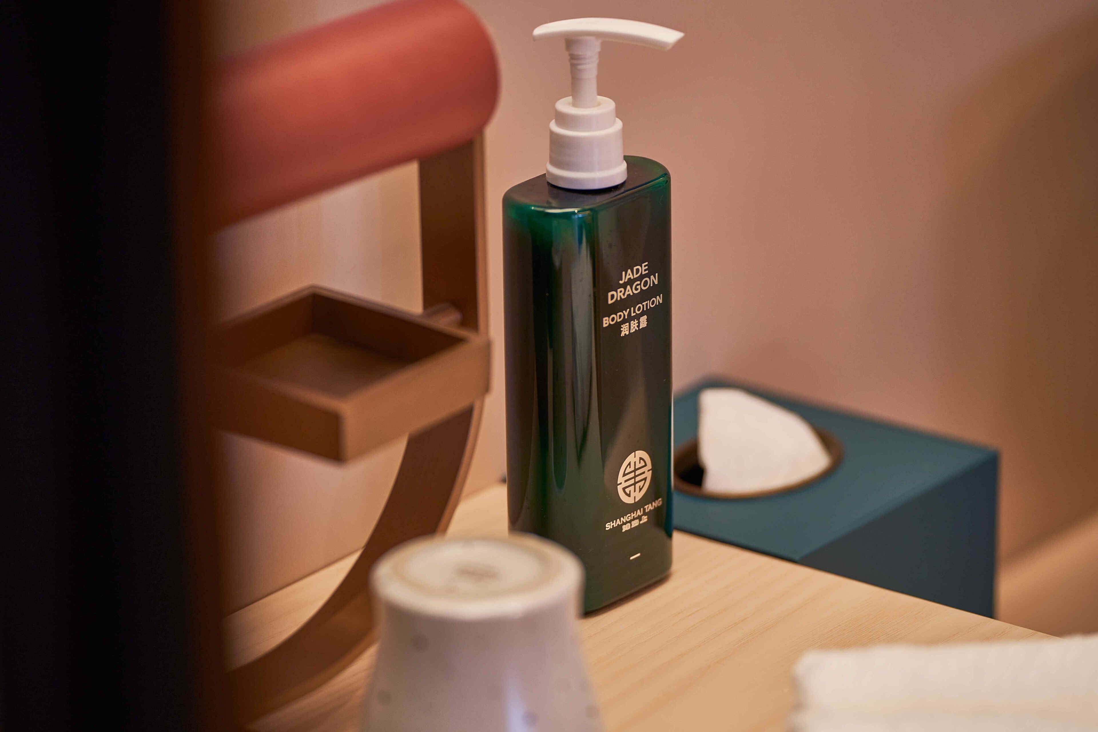 A bathroom with a green bottle of body lotion, a white pump dispenser, and a blue tissue box on a wooden surface.