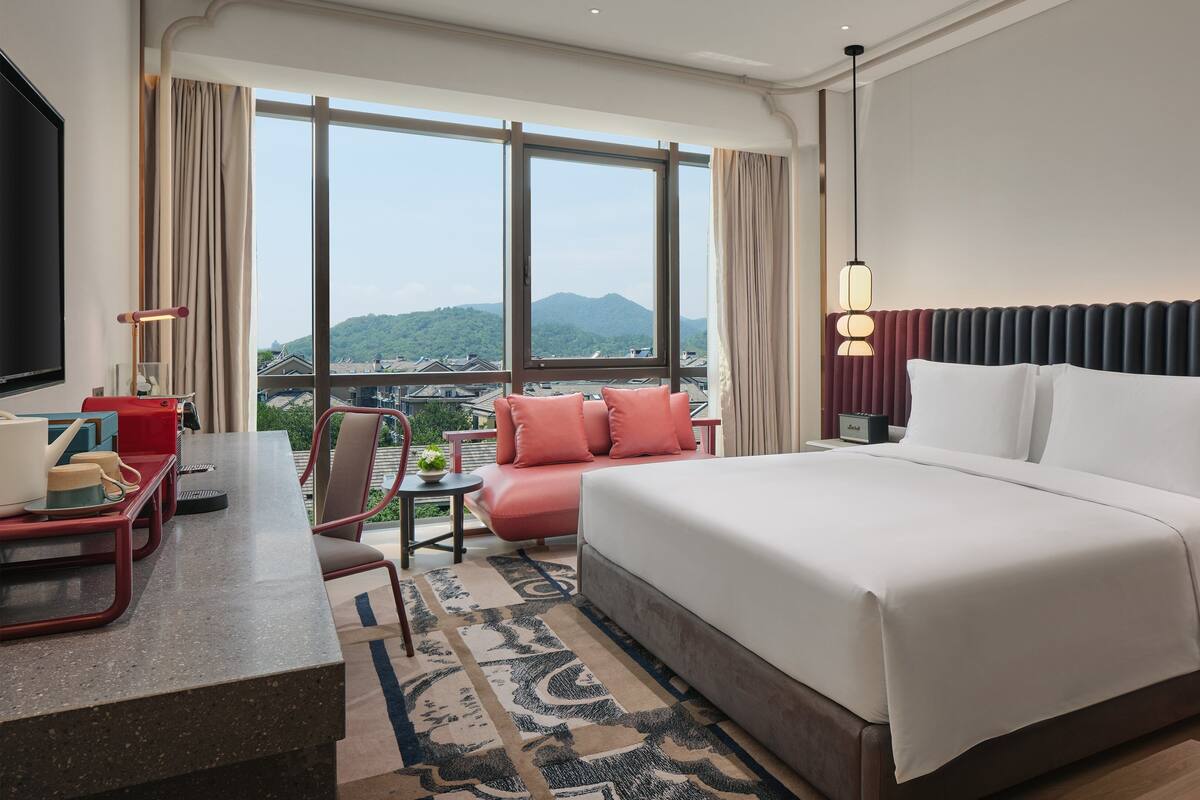 Ein Hotelzimmer mit einem großen Bett, einem Schreibtisch mit Stuhl, einem kleinen Tisch mit Lampe und Blick auf die Berge durch große Fenster.