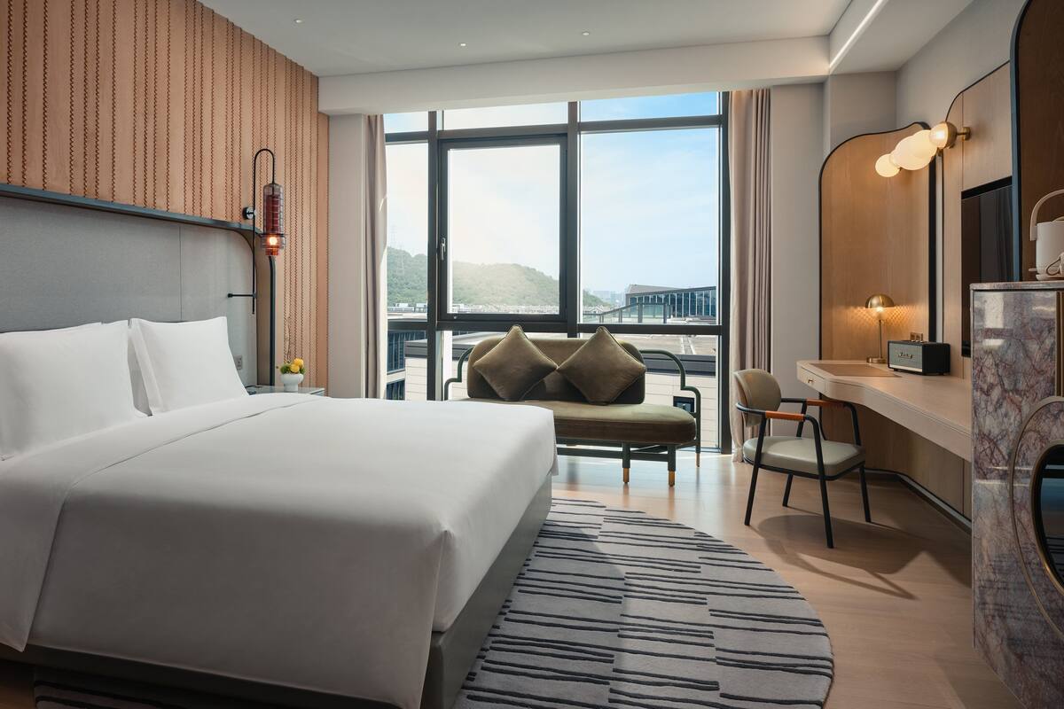 Ein modernes Hotelzimmer mit einem großen Bett, einer Sitzecke mit einer Couch und Sesseln, einem Schreibtisch mit einem Computer und einem großen Fenster mit Blick auf die Stadt.
