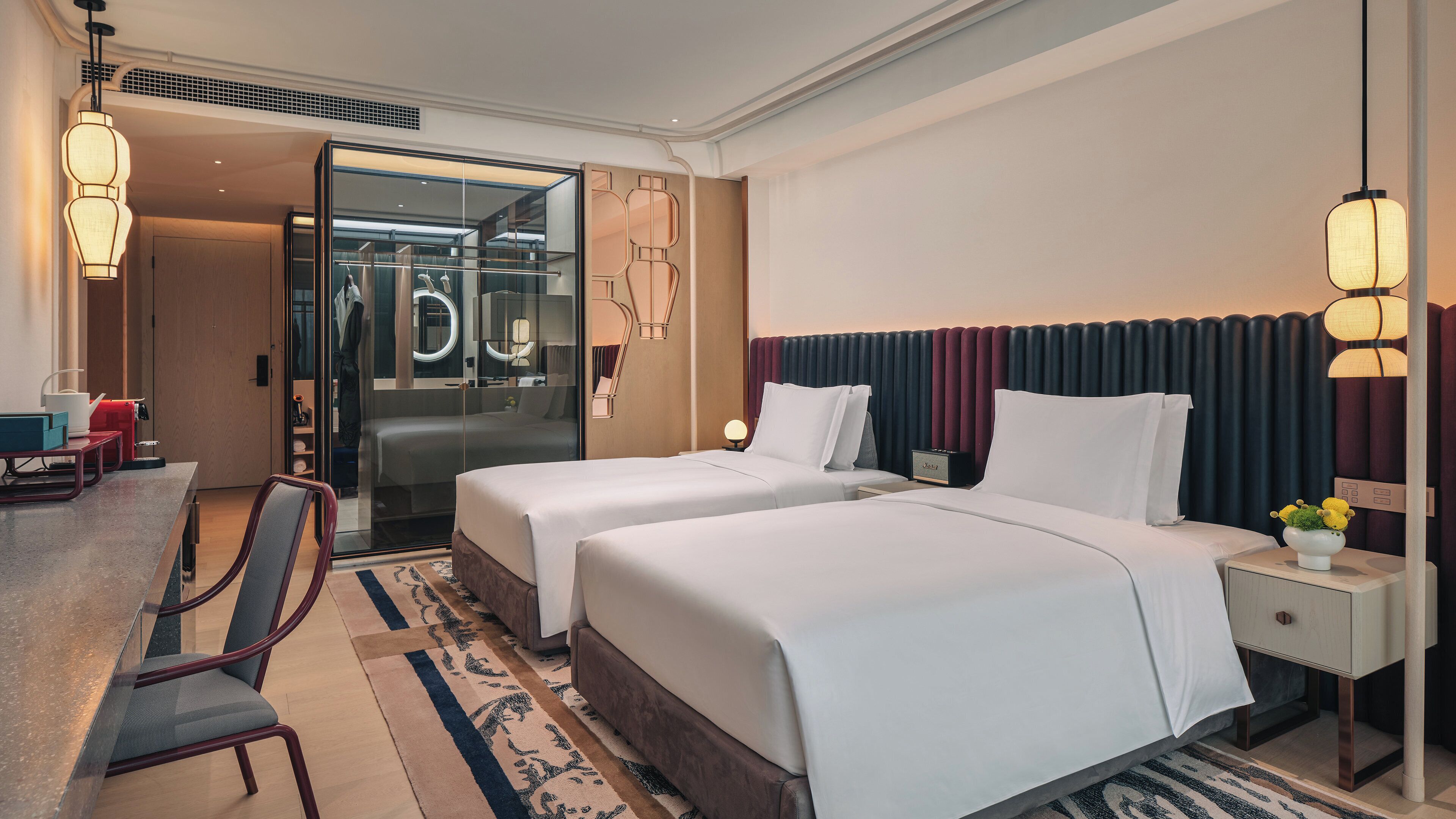A hotel room with two beds, a desk, a chair, a mirror, and a nightstand with a lamp.
