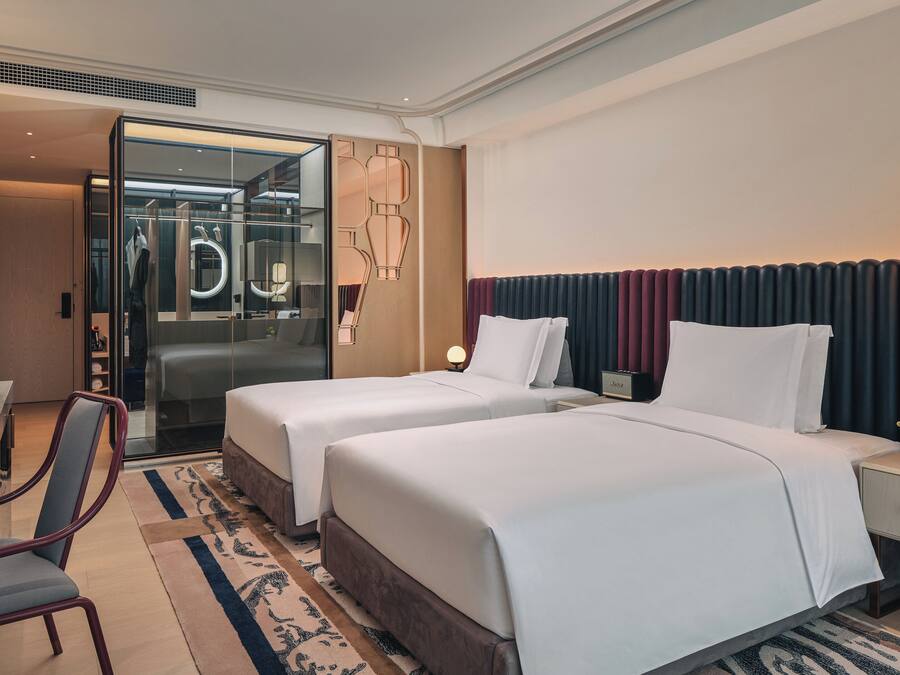 A hotel room with two beds, a desk, a chair, a mirror, and a nightstand with a lamp.