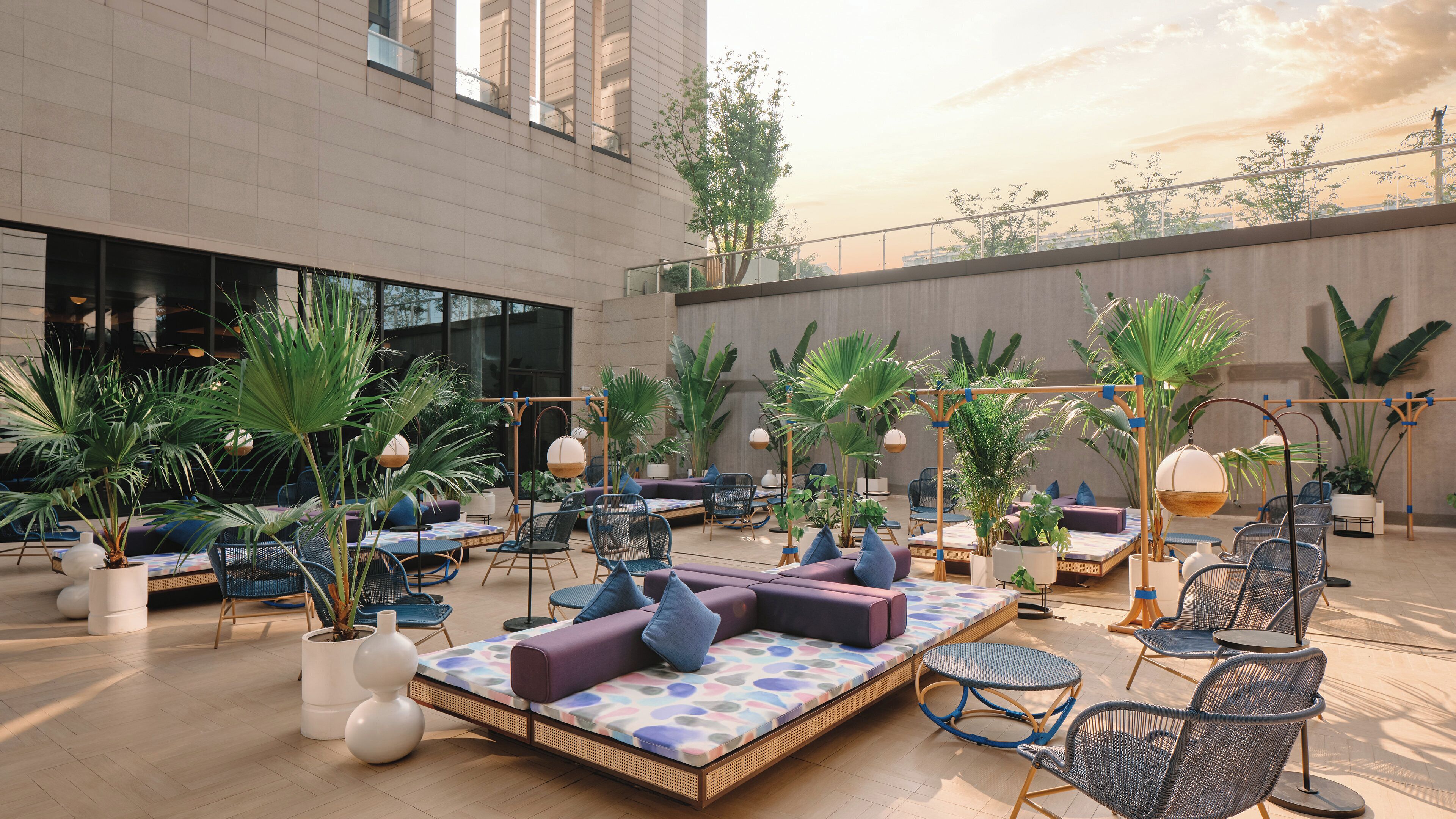 A spacious outdoor seating area with multiple lounge chairs and sofas, surrounded by potted plants and large windows.
