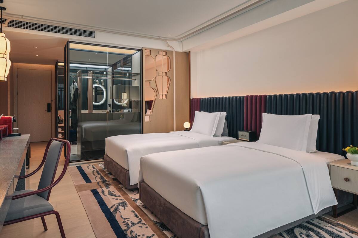 Ein Hotelzimmer mit zwei Betten, einem Schreibtisch, einem Stuhl, einem Spiegel und einem Nachttisch mit Lampe.