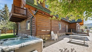 A log cabin with a hot tub, outdoor seating, and a stone fireplace.