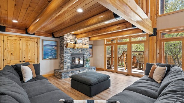 A spacious living room with a stone fireplace, wooden beams, and a large window overlooking a deck.