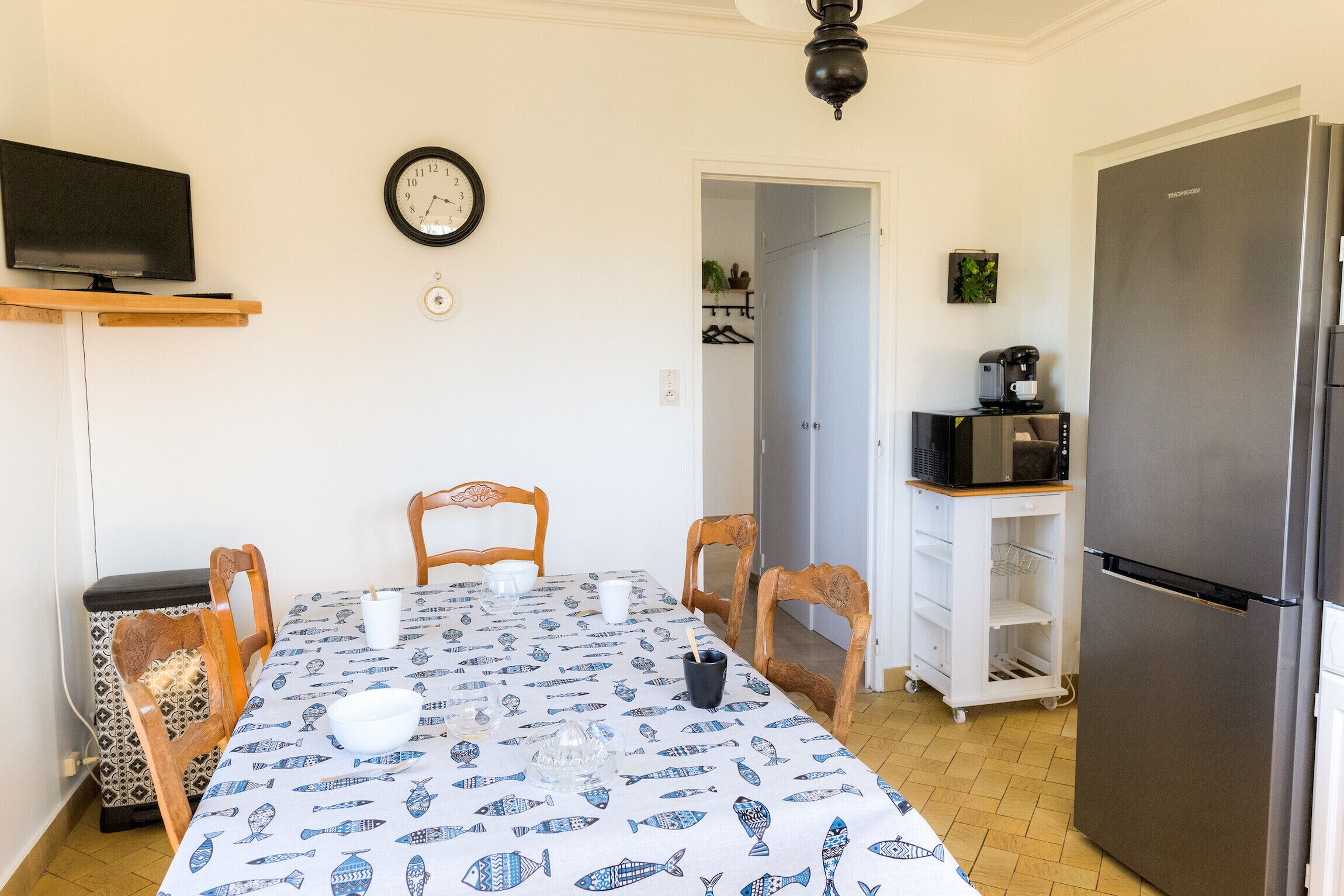 Un comedor con una mesa dispuesta para cuatro, un televisor arriba y un refrigerador a la derecha.