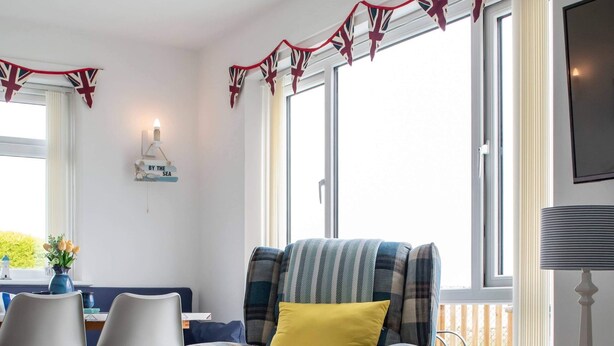 A cozy living room with a plaid armchair, a yellow pillow, and a black ottoman. There is a dining area with two chairs and a table, and a TV on a stand. - Stunning 3-bed Cottage in Lowestoft (Lowestoft)