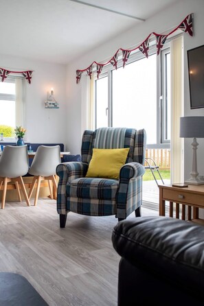 Un salon chaleureux avec un fauteuil à carreaux, un coussin jaune et un pouf noir. On y trouve également un coin repas avec deux chaises et une table, ainsi qu’un téléviseur posé sur un meuble.