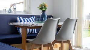 A dining area with a table, chairs, and a blue sofa. There is a window with curtains and a wall-mounted light fixture. - Stunning 3-bed Cottage in Lowestoft (Lowestoft)