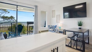 A modern kitchen with a large island, a dining area with a table and chairs, a balcony with chairs, and a flat-screen TV mounted on the wall. - Unmatched Views at Seaside Villa  Steps to the Sand (Hilton Head Island)
