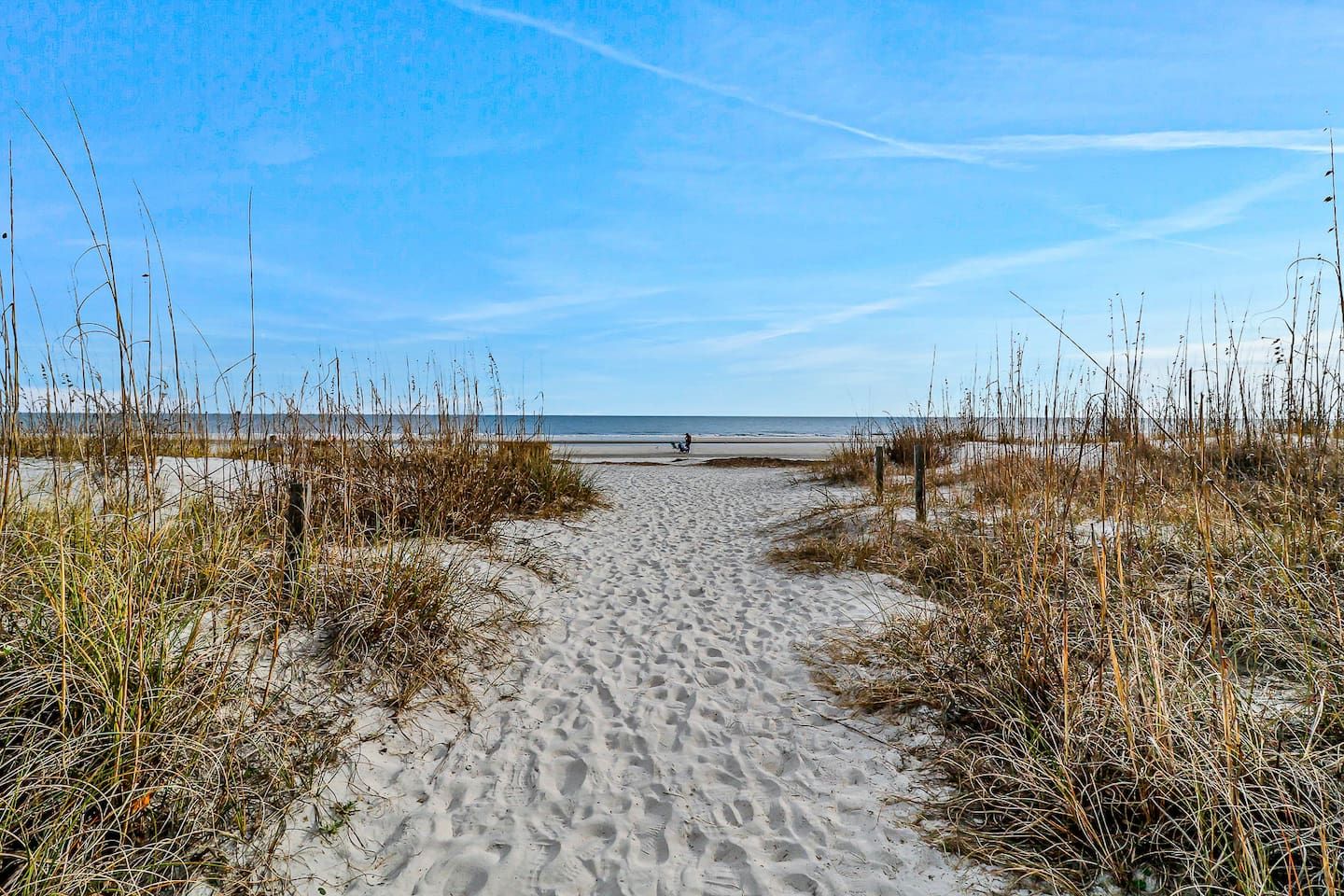 Ein Sandweg am Strand führt zum Meer, begleitet von spärlicher Vegetation an beiden Seiten.