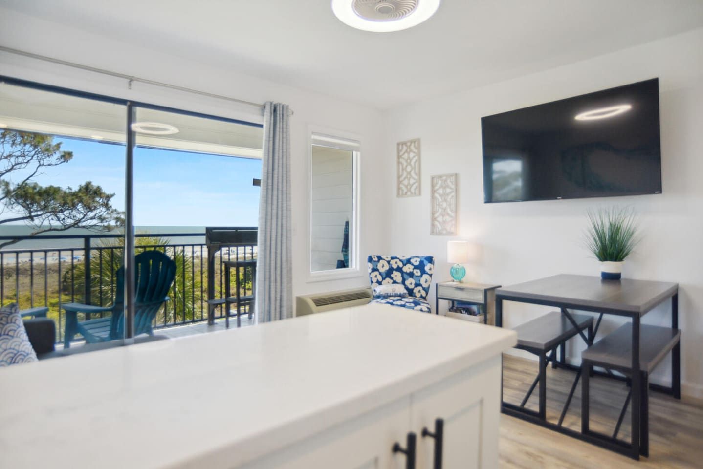 A modern hotel room with a balcony, a dining area, and a large flat-screen TV.