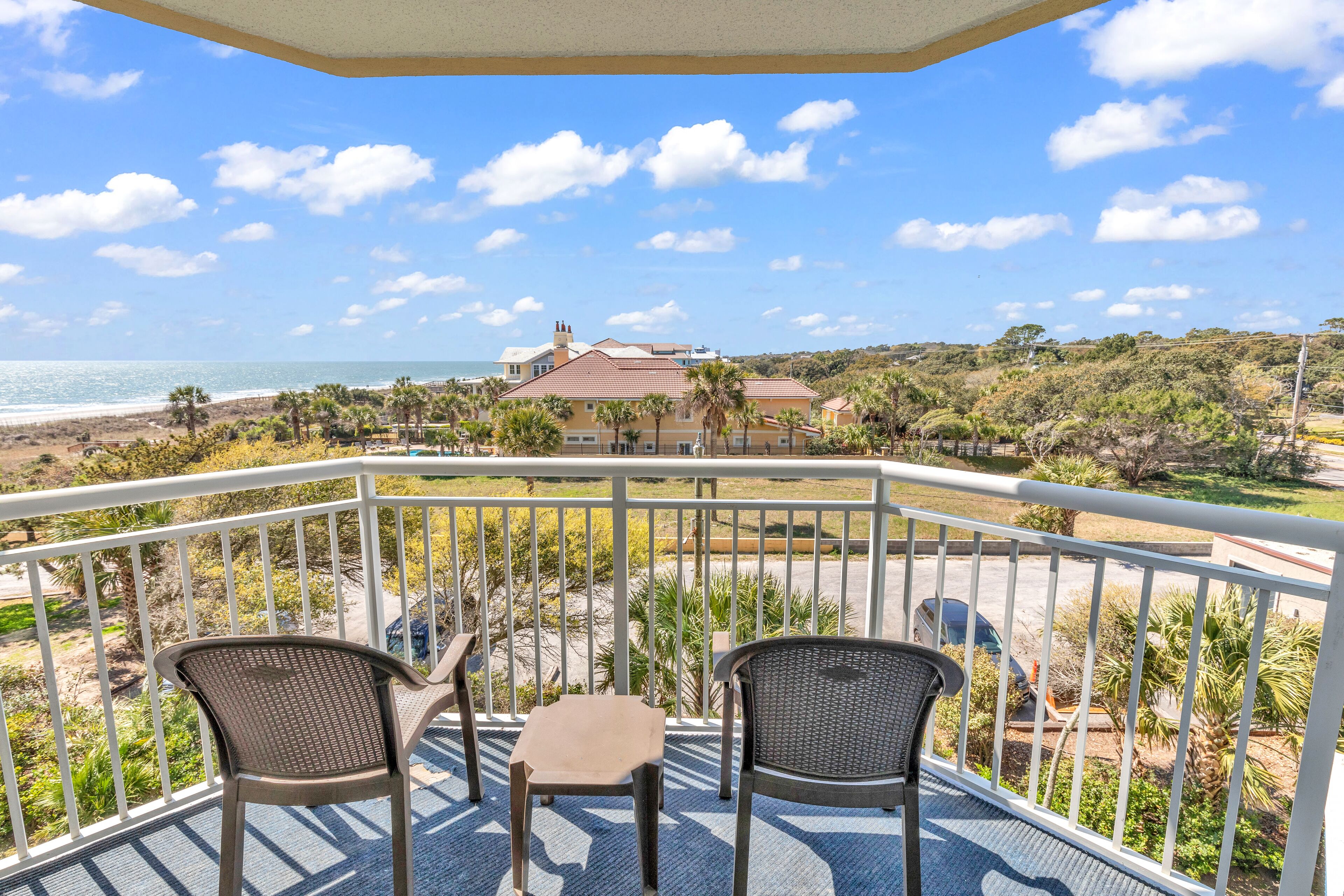 A balcony with two chairs and a table, overlooking a beach and a house.