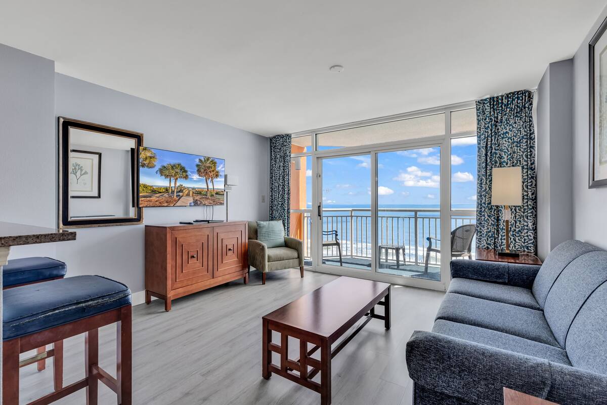 A modern living room with a sofa, a coffee table, and a cabinet. There is a large window with a view of the ocean.