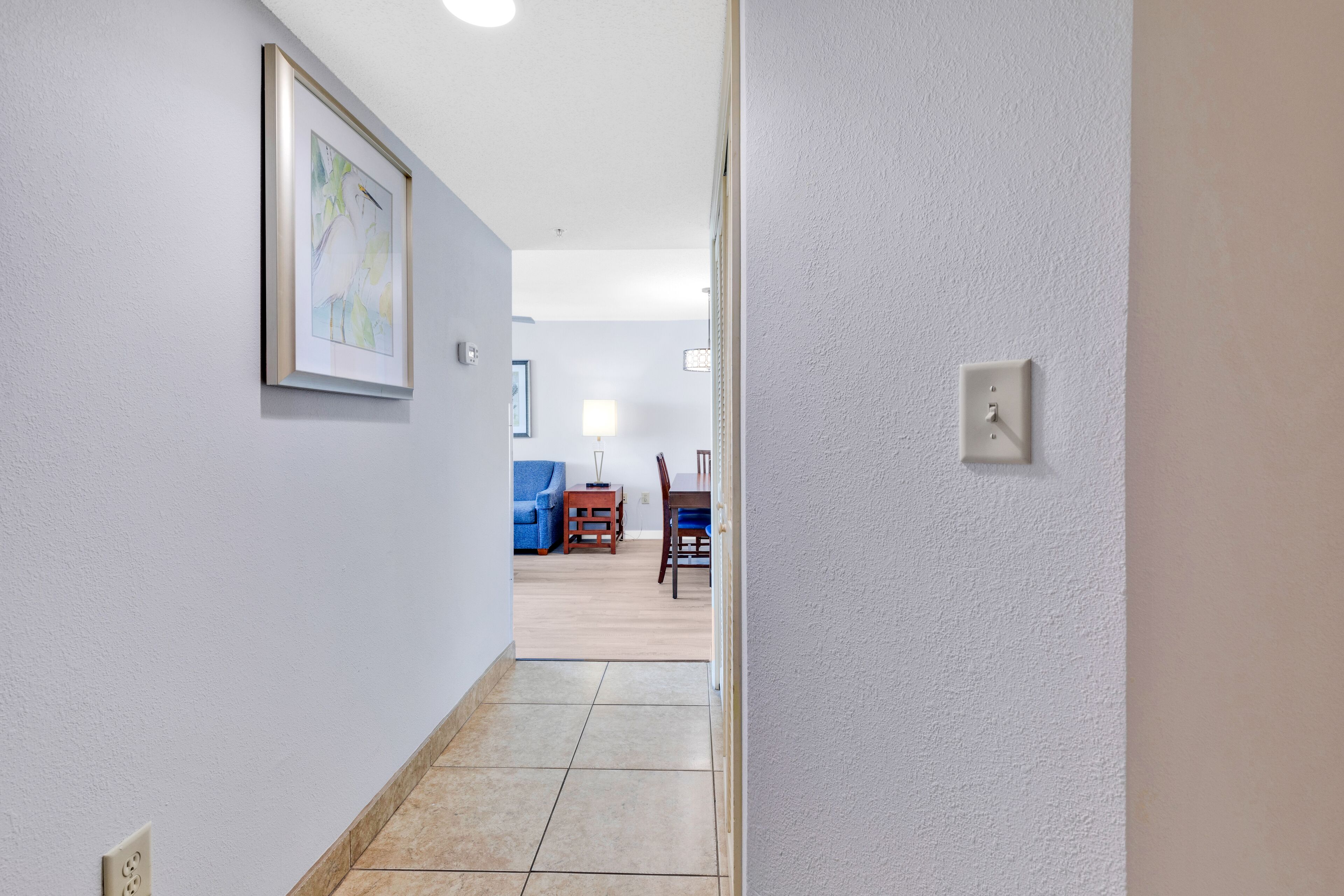 A hallway with a framed picture on the left, a dining area with a table and chairs in the background, and a light switch on the right wall.