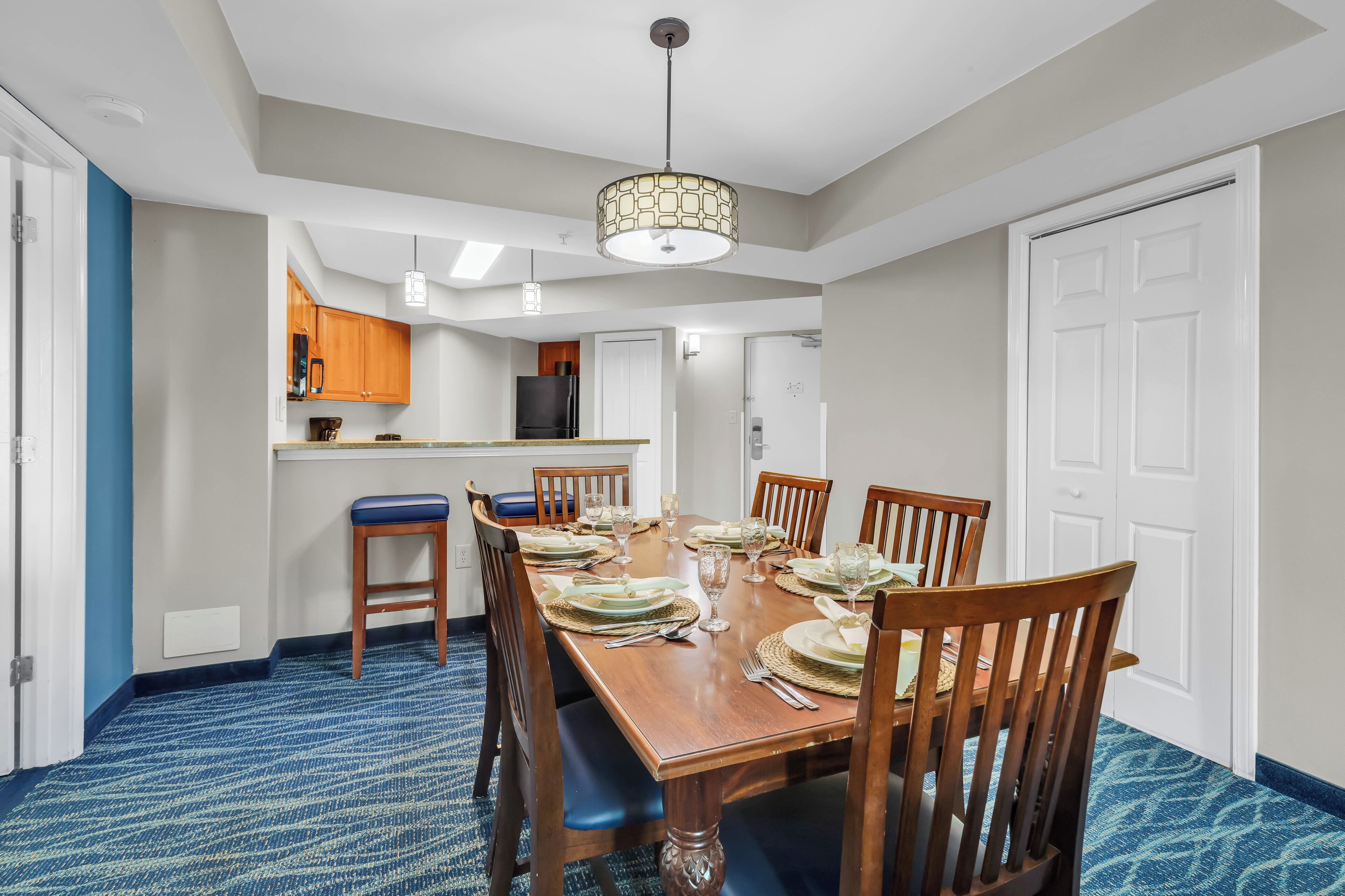 A dining area with a wooden table set for six, a kitchen in the background, and a blue carpeted floor.