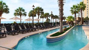 A pool area with lounge chairs and umbrellas, surrounded by palm trees and a view of the ocean. - Dunes Village Resort by Hosteeva (Myrtle Beach)