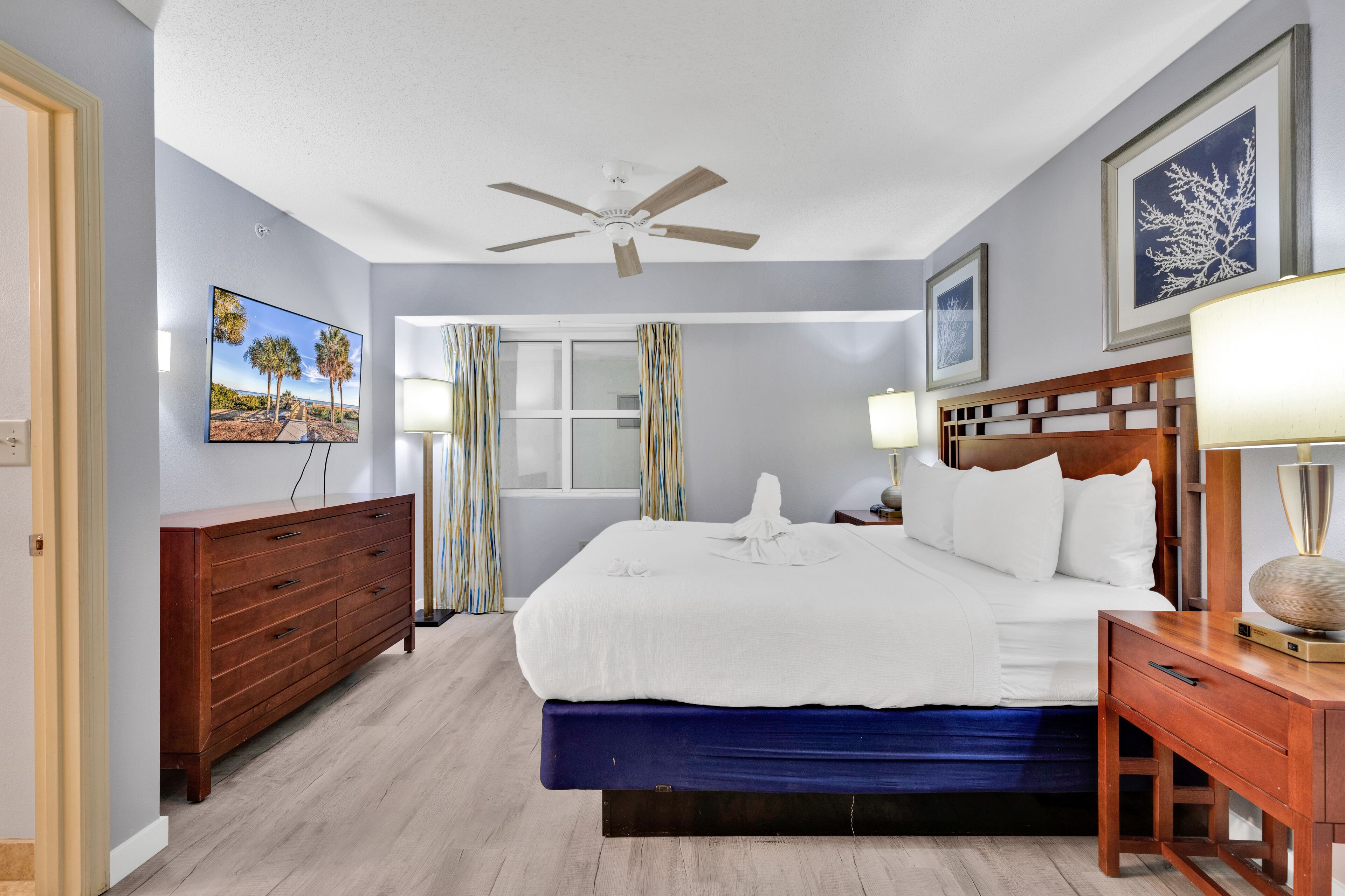 Une chambre d’hôtel avec un lit, une commode en bois, un ventilateur de plafond, une fenêtre avec des rideaux et un tableau au mur.