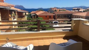 A balcony with two white chairs, a wooden fence, and a view of buildings and the sea. - The Broom (Golfo Aranci)