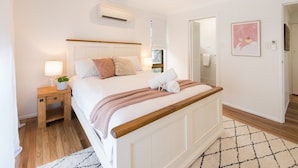 A bedroom with a bed, bedside table, lamp, and a wall-mounted air conditioner. - Beach House on Main- Airlie Beach (Airlie Beach)
