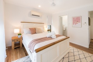 A bedroom with a bed, bedside table, lamp, and a wall-mounted air conditioner. - Beach House on Main- Airlie Beach (Airlie Beach)