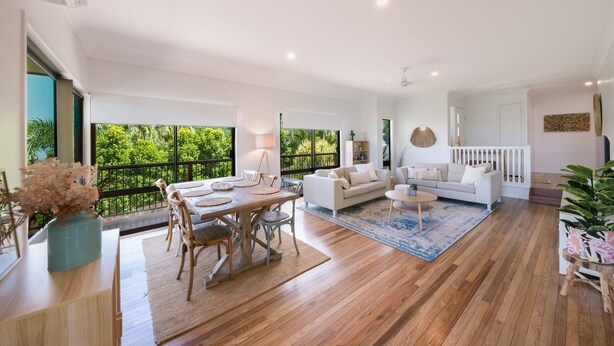A spacious living area with a wooden floor, a dining table with chairs, a white sofa, and a coffee table. There is a large window with a view of greenery outside.