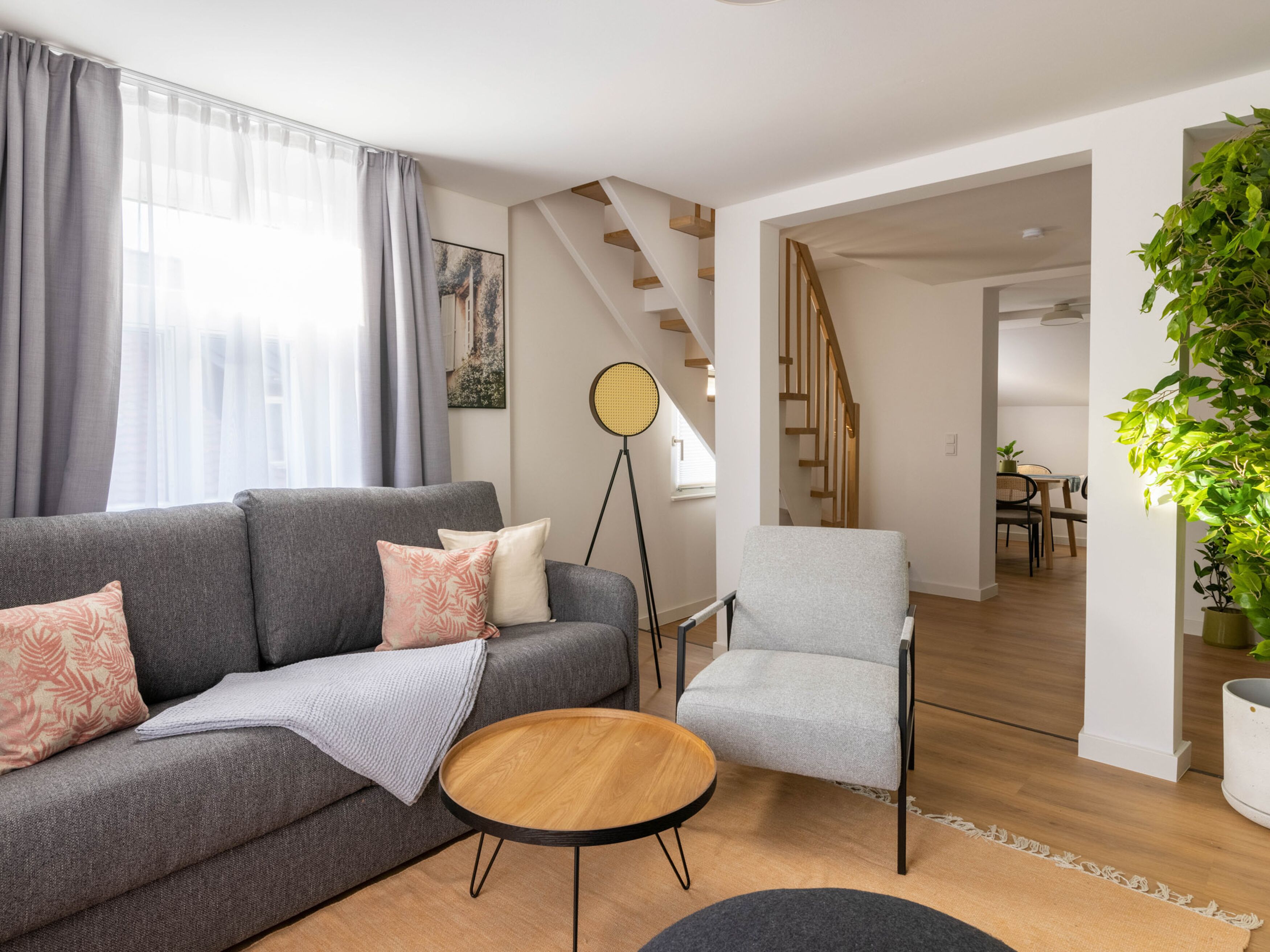 Un salon moderne avec un canapé gris, une table basse ronde en bois, un fauteuil gris et un lampadaire. Un escalier mène à une autre pièce.