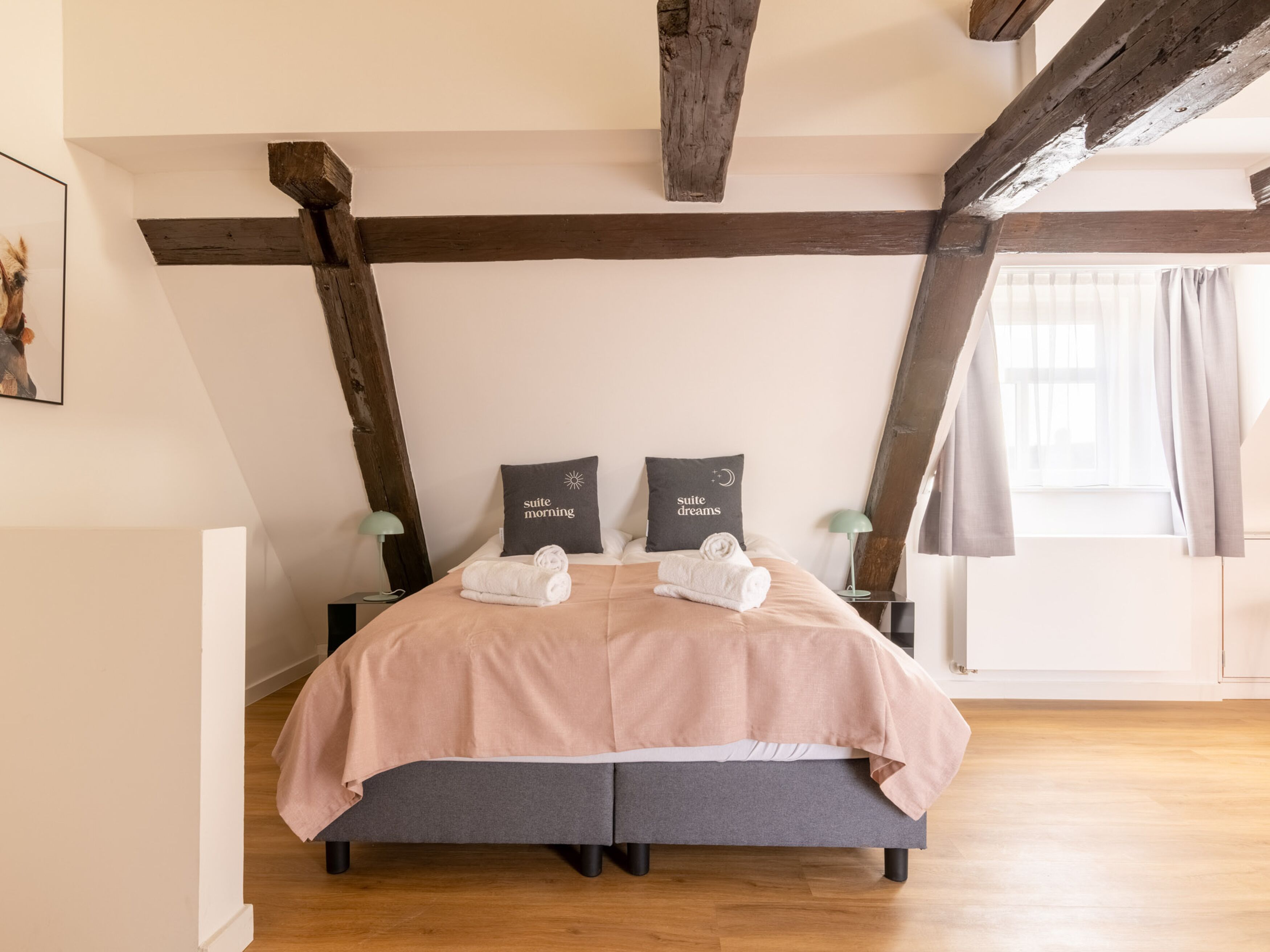 A bedroom with a bed, two bedside tables, a window with curtains, and exposed wooden beams on the ceiling.