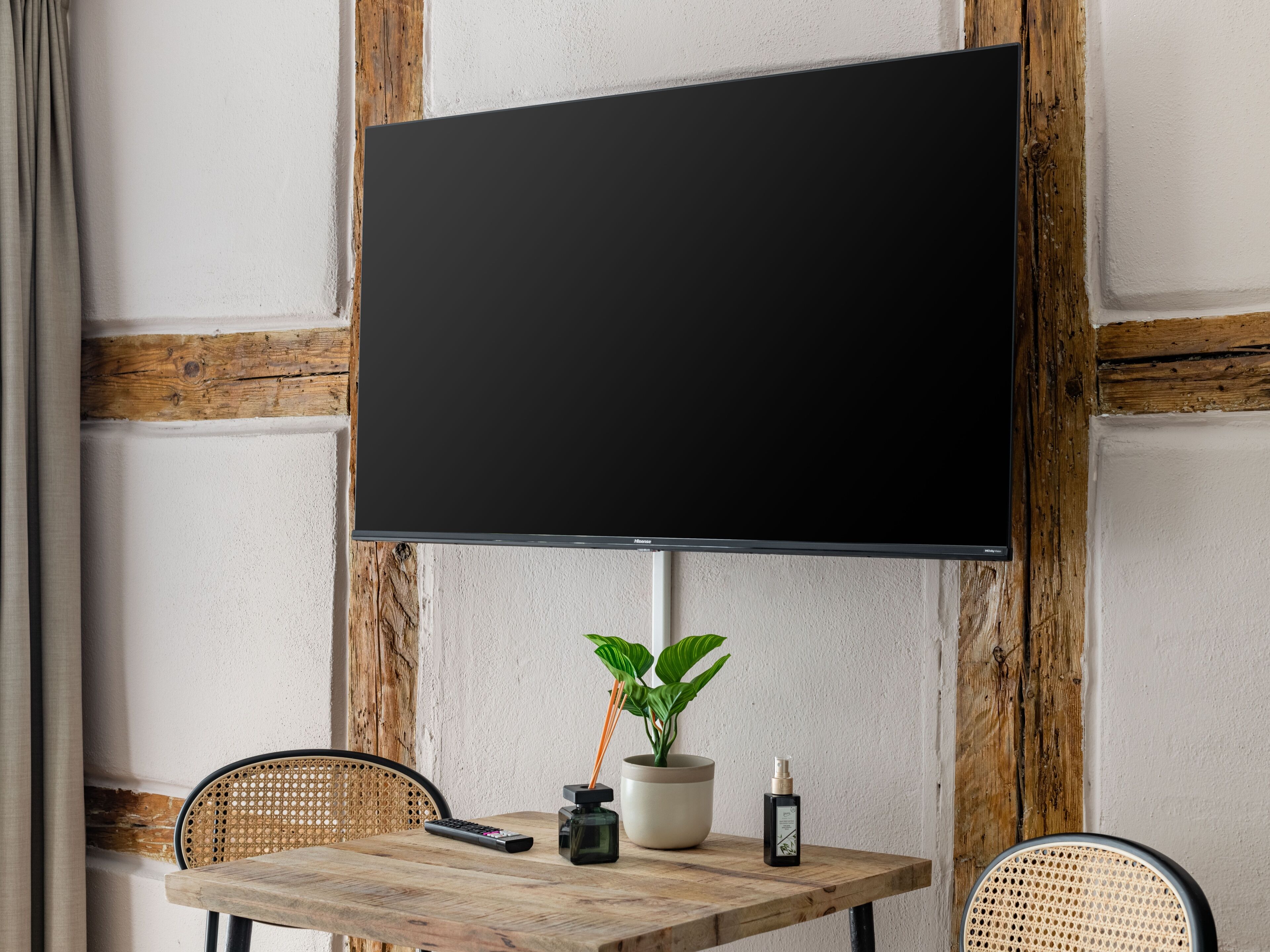 Een kamer met een grote, zwart tv-toestel aan een muur bevestigd, een houten tafel met een bloempot, een afstandsbediening en een kleine fles.